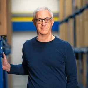 Andres Martin-Birner mit kurzen grauen haaren zwischen regalen im lager mit einem blauen pullover