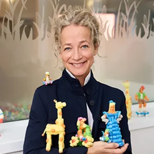 Business-Portr&auml;t von Britta Loick mit lockigem blondem Haar h&auml;lt bunte kreative Figuren aus Playmais in den H&auml;nden, umgeben von weiteren farbenfrohen Modellen.