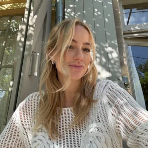 Business-Portrait von Christina Bischof mit blonden Haaren. Sie macht ein Selfie im Freien vor moderner Glasfassade, tr&auml;gt einen wei&szlig;en geh&auml;kelten Pullover und l&auml;chelt leicht.