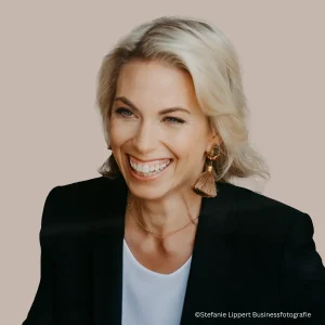 Business-Portr&auml;t von Dr. Jennifer Helgert lachend, blond Frau mit kurzem Bob in schwarzem Blazer und wei&szlig;em Shirt vor beigem Hintergrund