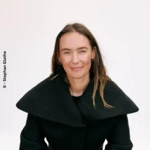 Business-Portr&auml;t von Jil Andert mit glattem braunem Haar in elegantem schwarzem Mantel mit breitem Kragen vor hellem Hintergrund