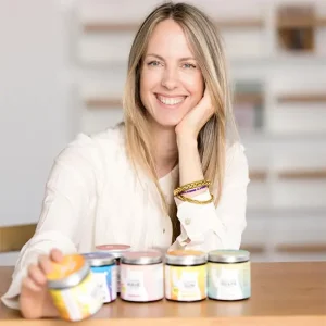 Business-Portr&auml;t von Laurence Saunier mit blonden, langen Haaren in heller Bluse, am Holztisch sitzend und pr&auml;sentiert mehrere bunte Lebensmittelgl&auml;ser.