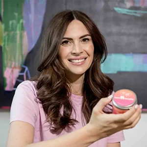 Business-Portr&auml;t von Marlena Hien mit langen, dunklen, welligen Haaren in einem rosa T-Shirt. Sie h&auml;lt ein kleines Glas mit Lebensmittelprodukt in die Kamera.