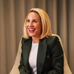 Business-Portr&auml;t von Mary-Jo C._Slipek mit blondem Bob in dunkelgr&uuml;nem Blazer und wei&szlig;em Shirt sitzt vor beige-grauem Vorhang, professionelles Business-Portr&auml;t mit moderner, energiegeladener Ausstrahlung