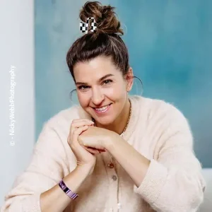 Business-Portr&auml;t von Stefanie Lischka mit lockerem Dutt und kariertem Haar-Clip, tr&auml;gt eine helle Strickjacke und sitzt entspannt vor blauem Hintergrund