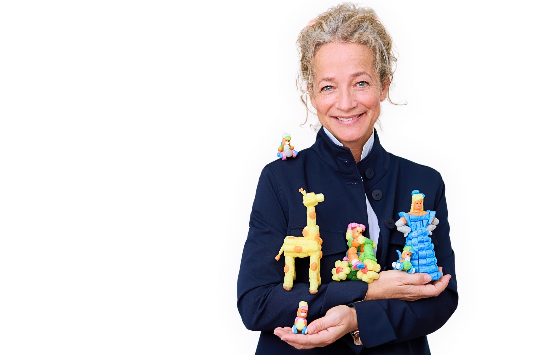 Freigestelltes Porträt von Britta Loick, CEO von Playmais, lächelnd in dunkelblauer Jacke. Sie hält mehrere farbenfrohe Playmais-Figuren in den Händen, darunter eine Giraffe, eine Prinzessin und weitere kleine Figuren. Heller Hintergrund mit dezentem, mehrfarbigem Outline-Effekt um die Person.