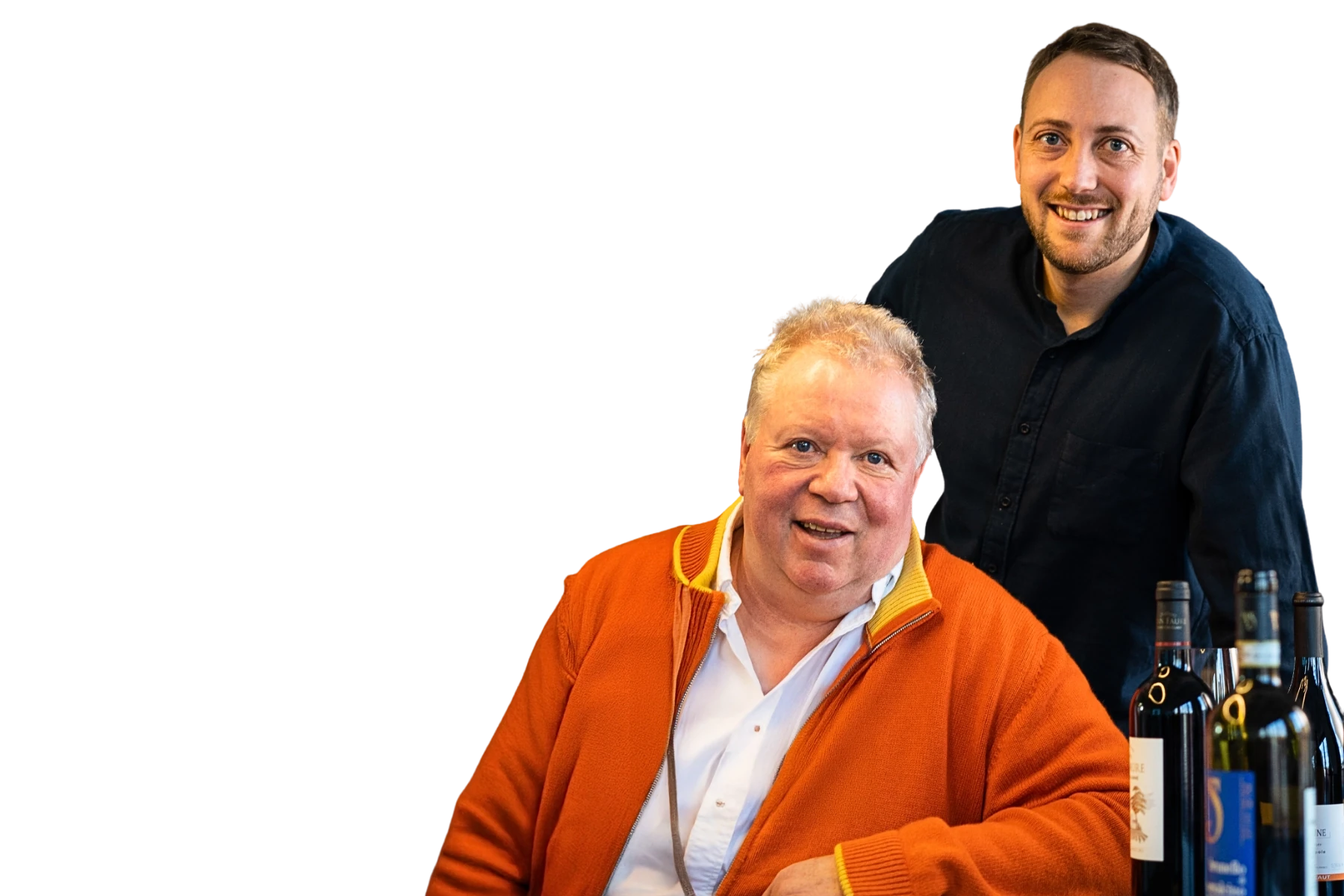 Freigestelltes Porträt von Heiner Lobenberg (sitzend, vorne) und Luca Lobenberg (stehend, hinten) von Lobenberg’s Gute Weine. Beide lächeln in die Kamera. Heiner trägt einen orangefarbenen Cardigan über einem weißen Hemd, Luca ein dunkles Hemd. Rechts im Bild stehen mehrere Weinflaschen. Freisteller zur K5 ChefTreff Podcast Episode 269.