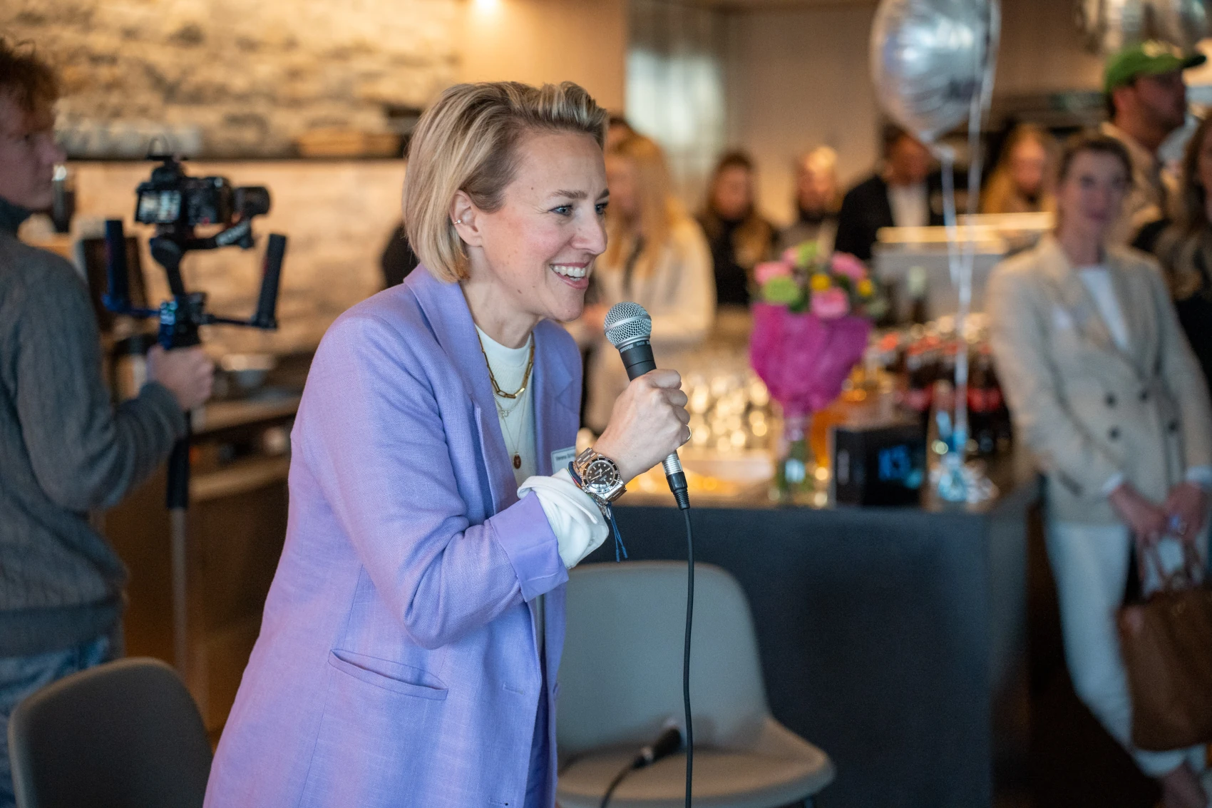 Verena Schlüpmann, mit blonden kurzen Haaren in hellblauem Blazer am Mikrofon während eines K5 MEET UP Events, lachend vor Publikum in warmer Restaurant-Atmosphäre mit Kamerateam im Hintergrund.
