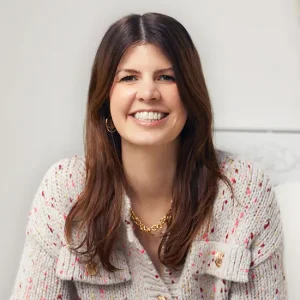 Alexandra Szarmach l&auml;chelnd mit langen braunen Haaren in gemusterter Tweedjacke und goldener Kette vor hellem Hintergrund.