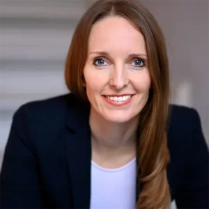 Anna Kaiser freundlich l&auml;chelnd mit langen braunen Haaren und blauen Augen im dunklen Blazer und hellem Shirt