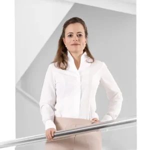 Professionelles Business-Portr&auml;t von Bonita Grupp in wei&szlig;er Bluse und beiger Hose, selbstbewusst stehend an einem Gel&auml;nder vor moderner Architektur