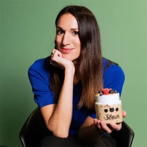 L&auml;chelnde Caroline Nichols mit langen dunklen Haaren in blauem Oberteil h&auml;lt ein Dessertglas mit Beeren in der Hand vor gr&uuml;nem Hintergrund