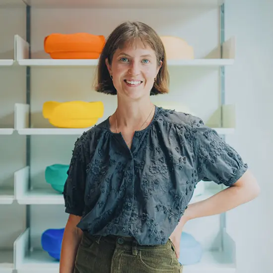 Hannah K&ouml;nig l&auml;chelnd mit kurzem braunem Bob in strukturierter Bluse steht vor Regal mit farbigen Design- oder Produktobjekten
