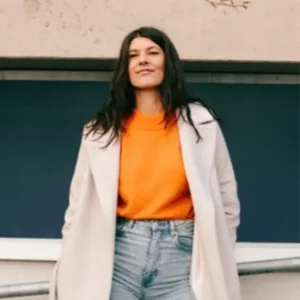 Lena Jüngst mit langen dunklen Haaren tr&auml;gt orangefarbenen Pullover, hellen Mantel und Jeans, steht im urbanen Au&szlig;enbereich vor dunkler Wand