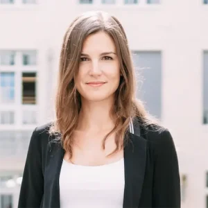 Professionelles Portr&auml;t von Lenia Karallus mit schulterlangen braunen Haaren in schwarzem Blazer und wei&szlig;em Top vor urbanem, hellem Hintergrund