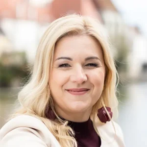 Petra Gruber l&auml;chelnd in hellem Blazer und bordeauxfarbenem Oberteil vor urbanem Hintergrund mit Wasser, nat&uuml;rliches Business-Portr&auml;t f&uuml;r Beratung, Coaching und Personal Branding