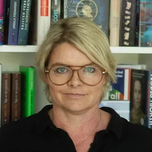 Portr&auml;t von Sarah Orlandi mit kurzem blondem Haar und runder Brille vor B&uuml;cherregal, tr&auml;gt schwarzes Oberteil