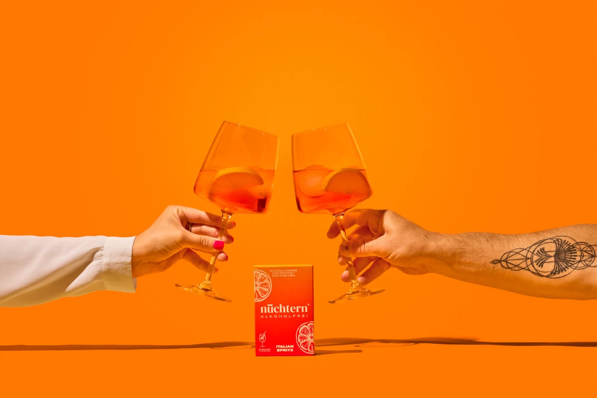 Lifestyle-Thumbnail: Zwei Hände stoßen mit Spritz-Gläsern an, im Zentrum steht eine rote Packung „nüchtern ALKOHOLFREI Italian Spritz“ auf orangem Hintergrund.