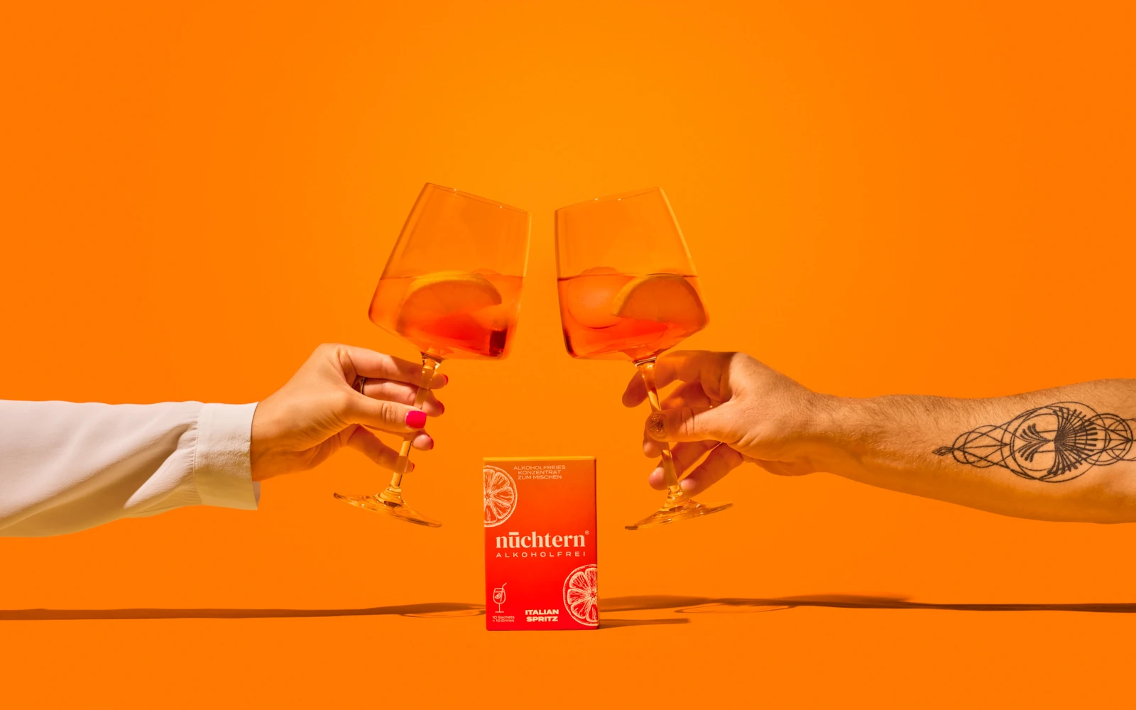 Zwei Personen stoßen mit leuchtend orangen, alkoholfreien Drinks an. Im Vordergrund steht eine Packung "nüchtern. alkoholfei - ITALIAN SPRITZ". Das Bild ist vor einem knalligen orangen Hintergrund inszeniert und symbolisiert das "Mindful Drinking" Konzept von nuechtern.berlin beim K5 Event.