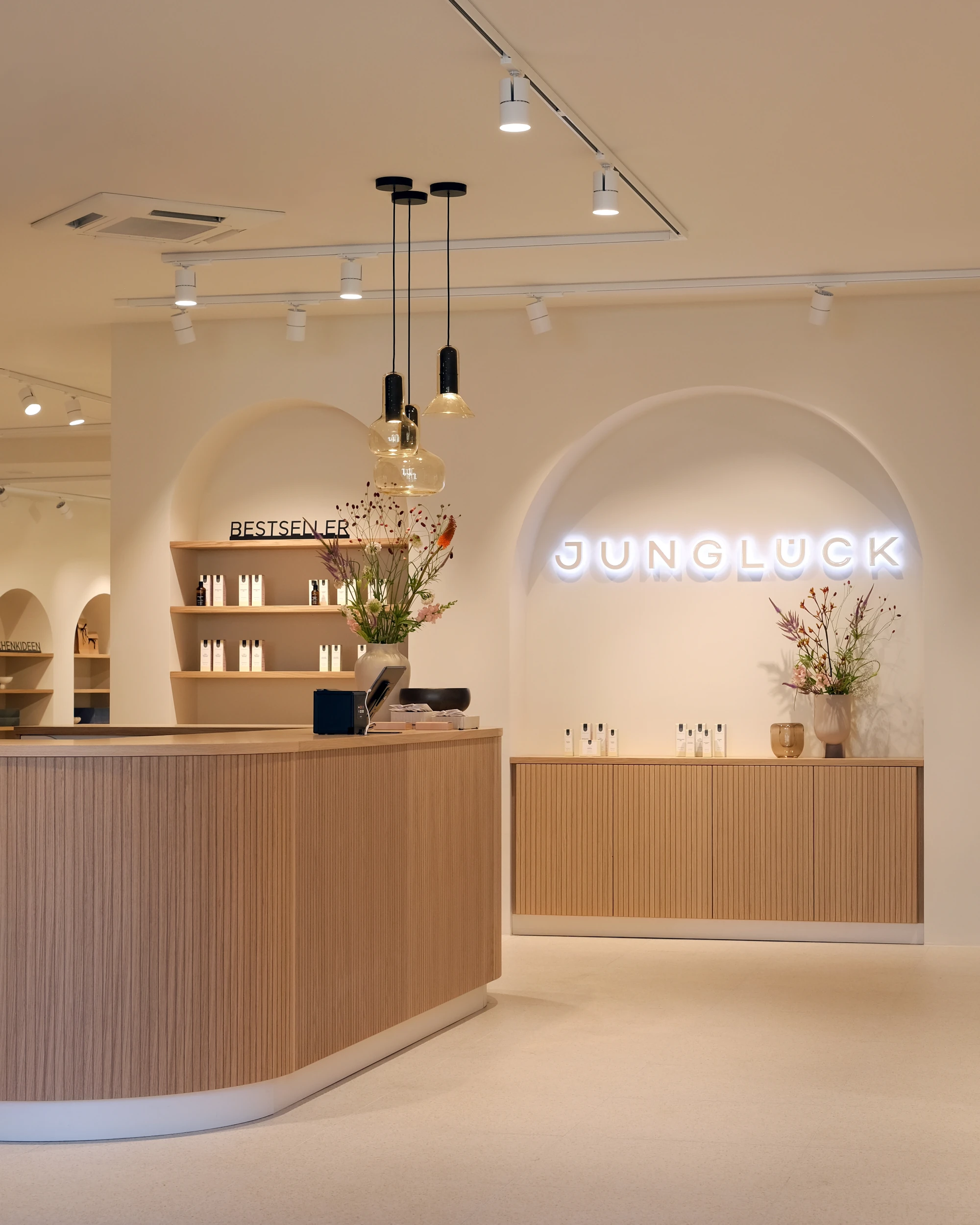 Innenansicht des Junglück Flagship Stores in München am Gärtnerplatz. Zu sehen sind der hölzerne Kassentresen, das beleuchtete 'JUNGLÜCK'-Neonlogo in einer Wandnische sowie Regale mit Kosmetikprodukten. Location-Bild für das K5 Female Circle Meetup.