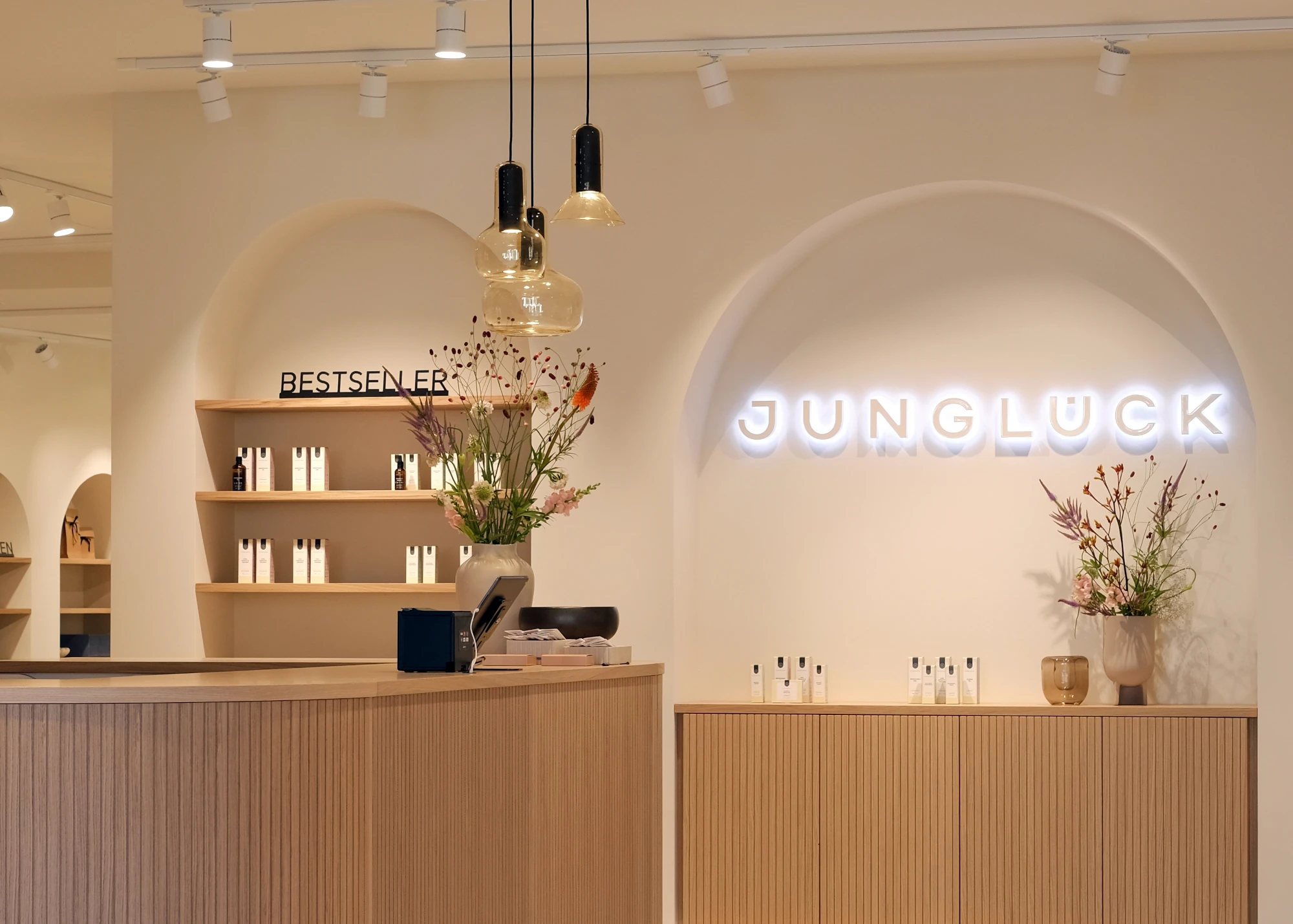 Interieur des Junglück Flagship Stores in München am Gärtnerplatz. Zu sehen sind der hölzerne Kassentresen mit Lamellenstruktur, das beleuchtete weiße 'JUNGLÜCK'-Neonlogo an einer beigen Bogenwand sowie Regale mit Hautpflegeprodukten und Blumenarrangements.