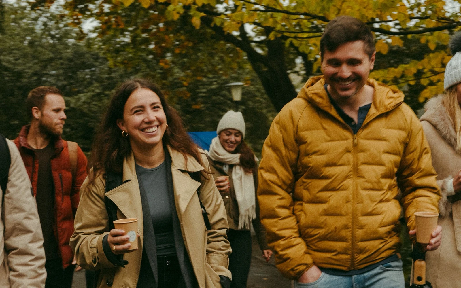 Lächelnde Teilnehmerinnen und Teilnehmer beim K5 Female Circle Recovery Walk by LOOSH. Die Gruppe spaziert in herbstlicher Atmosphäre im Freien, hält Kaffeebecher in der Hand und unterhält sich angeregt.