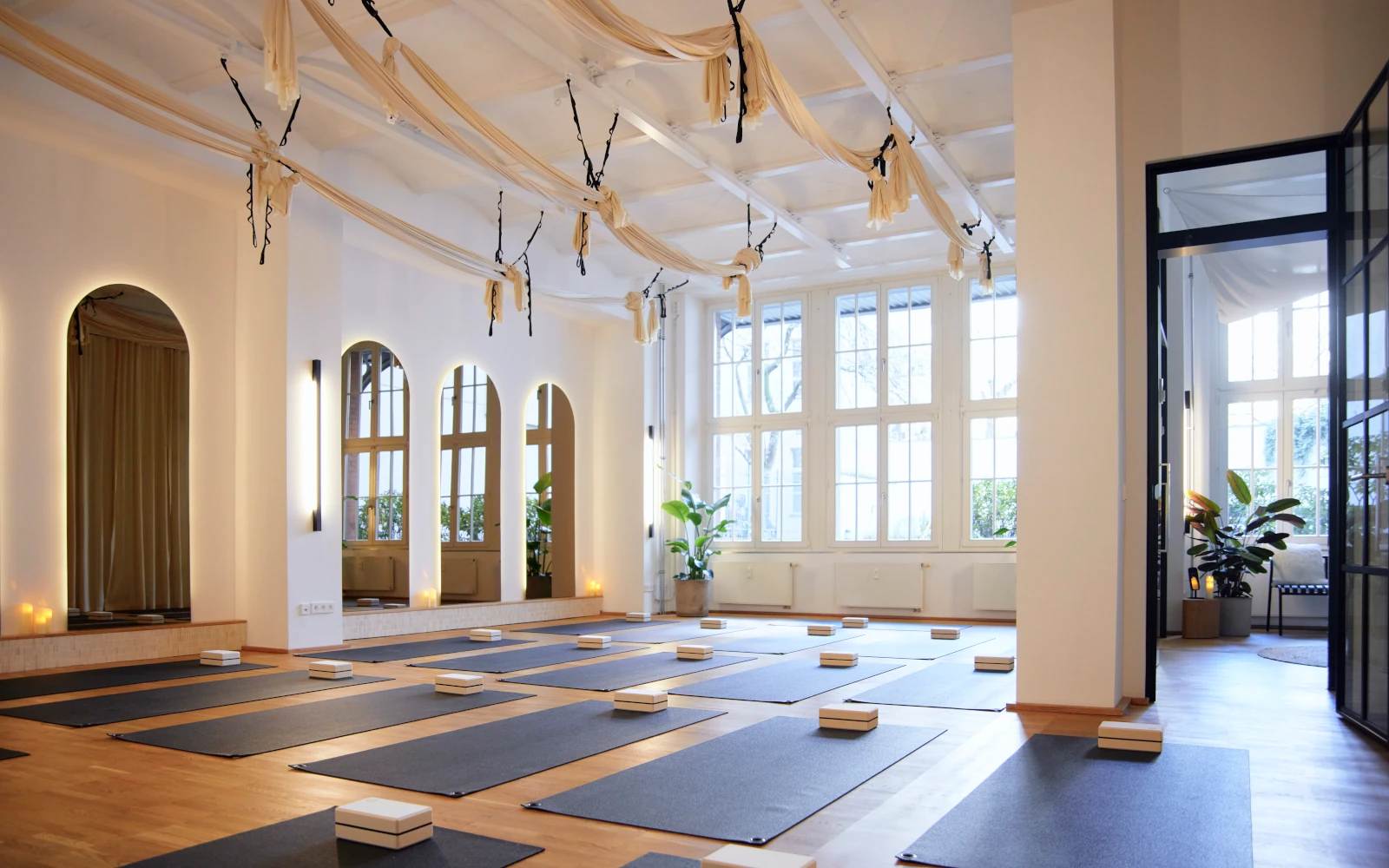 Heller, lichtdurchfluteter Übungsraum im Hale.Now Studio mit schwarzen Pilates-Matten, Blöcken und großen Bogenfenstern. Von der Decke hängen helle Tücher, und warme Beleuchtung schafft eine einladende Atmosphäre für den Pilates-Kurs.
