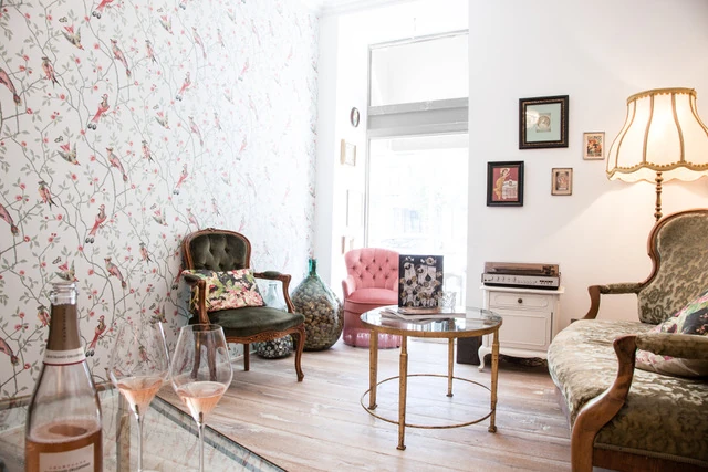 Gemütliches, stilvolles Interieur der Champagne Boutique in München. Vor einer floralen Tapete mit Vogelmotiven stehen antike Samtsessel in Grün und Rosa um einen runden Glastisch, auf dem Rosé-Champagner bereitsteht.