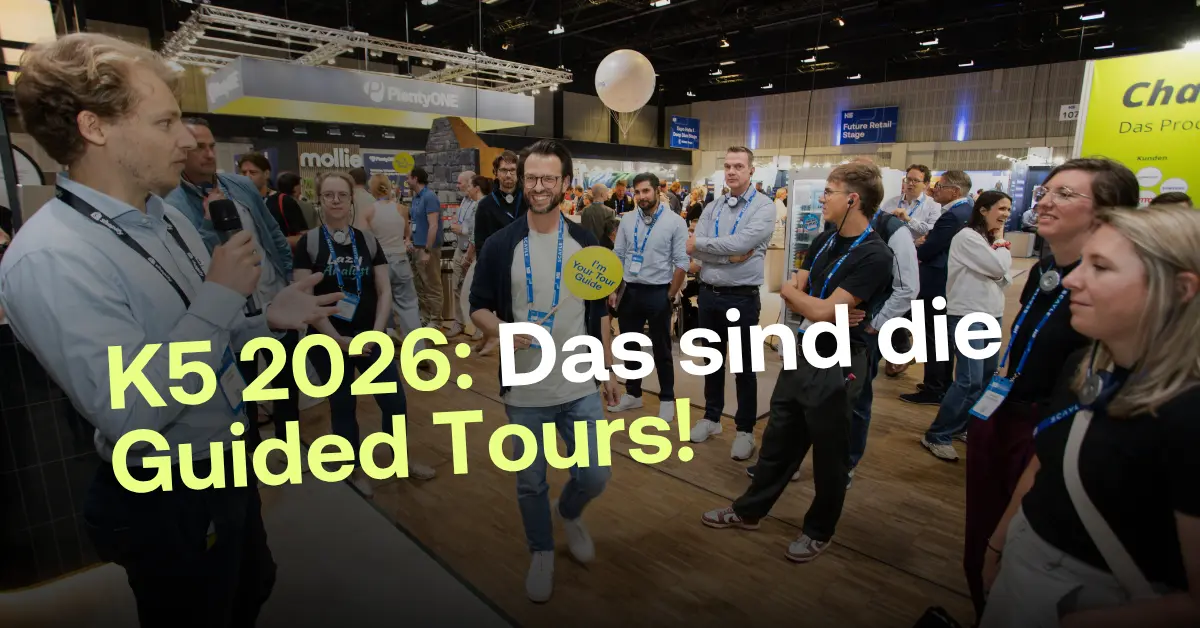 Eine Gruppe von Menschen auf der K5 Konferenz 2026. Ein Tourguide mit einem gelben Schild &bdquo;I'm Your Tour Guide&ldquo; l&auml;chelt in die Kamera, w&auml;hrend im Vordergrund ein Sprecher mit Mikrofon eine gef&uuml;hrte Tour leitet.