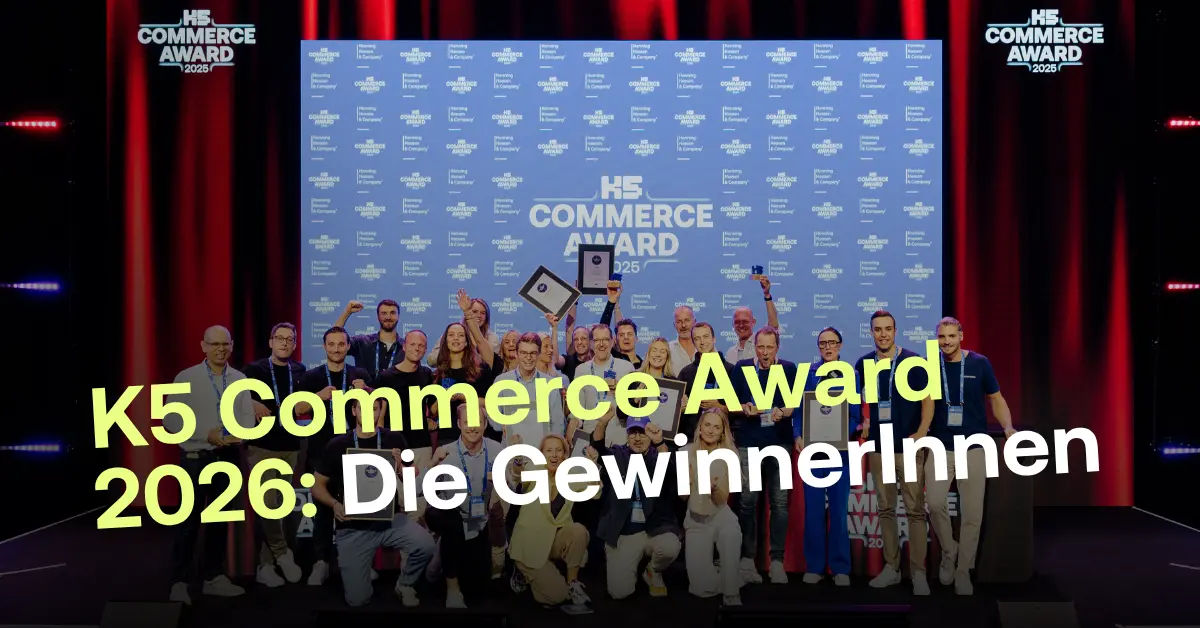 Eine gro&szlig;e Gruppe von jubelnden Gewinnerinnen und Gewinnern steht mit Pokalen und Urkunden auf einer Eventb&uuml;hne vor einer blauen Sponsorenwand des K5 Commerce Awards.