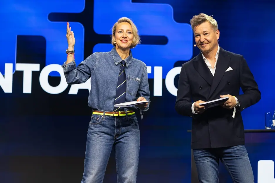 Verena Schlüpmann und Sven Rittau moderieren auf der K5 Bühne. Die Frau, links, trägt ein Jeanshemd, Jeans, eine Krawatte und einen gelben Gürtel und hebt die rechte Hand. Der Mann, rechts, trägt einen dunklen Doppelreiher-Anzug über einem weißen Hemd und Jeans und hält Unterlagen. Beide lächeln vor einem großen blauen Hintergrund mit weißer Schrift.