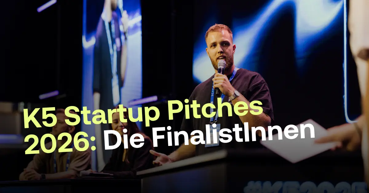 Ein junger Mann mit Bart spricht auf einer Event-B&uuml;hne konzentriert in ein Mikrofon w&auml;hrend der K5 Startup Pitches 2026. Im Hintergrund sitzen weitere Personen vor einer gro&szlig;en, blau leuchtenden LED-Wand.